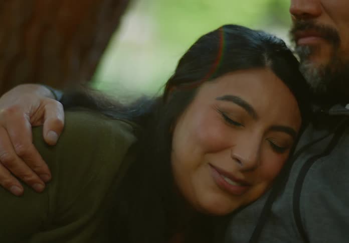 Older couple smiles and talks while man hugs and kisses woman as they sit near tree