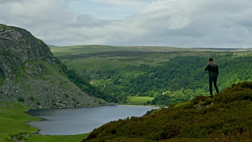 A man on a cell phone stands looks down over a green valley with a lake