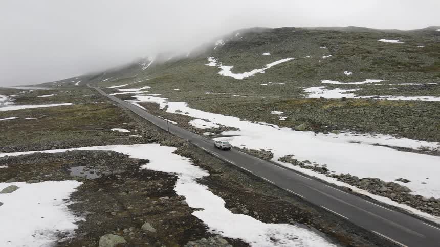Aerial panorama of a car traveling on a road at the edge of snowy mountains on a cloudy day