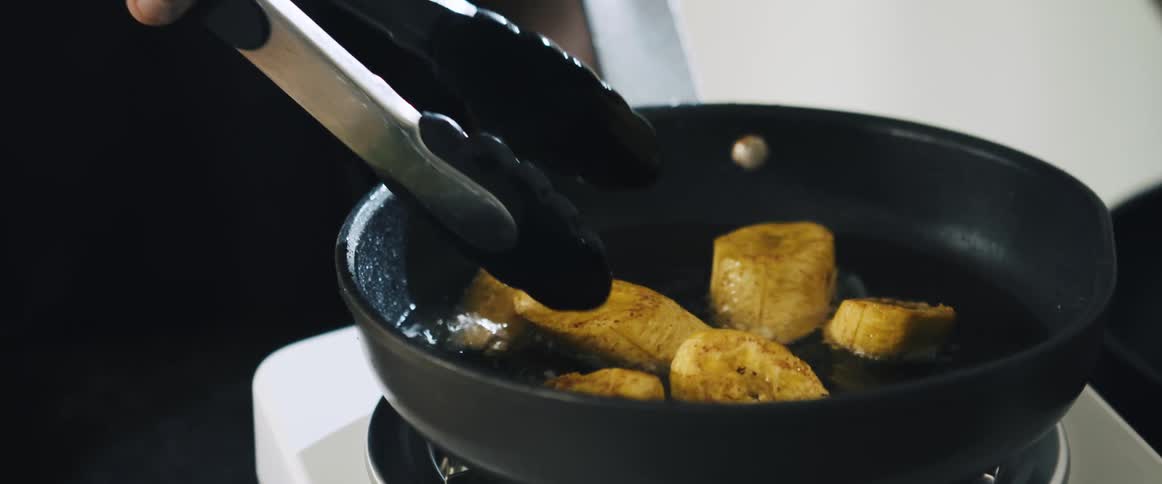 Person with a spatula frying plantain slices in a frying pan with oil