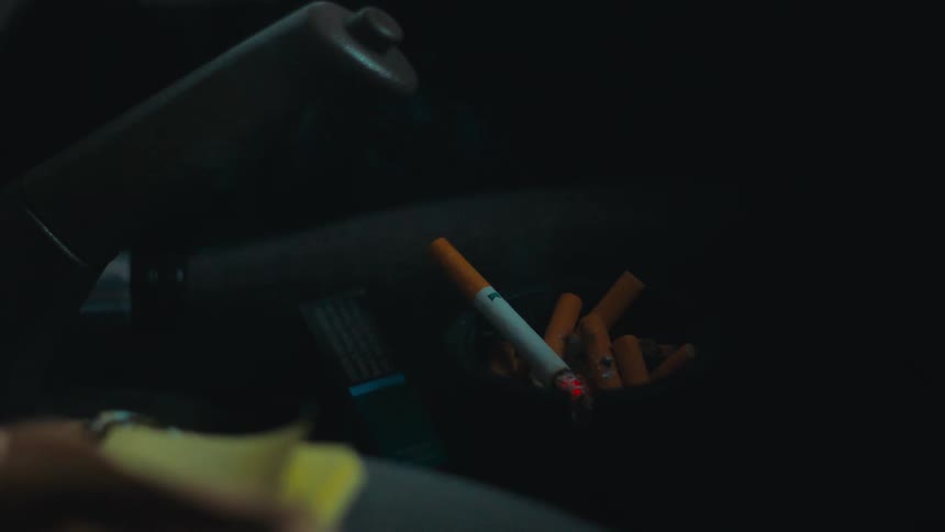 A hand in a car laying a lit cigarette on a full ashtray near the handbrake