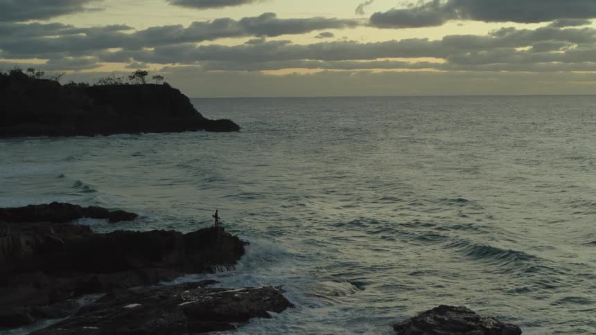 Silhouette of a person holding a surfboard on a seaside rock in a morning sky