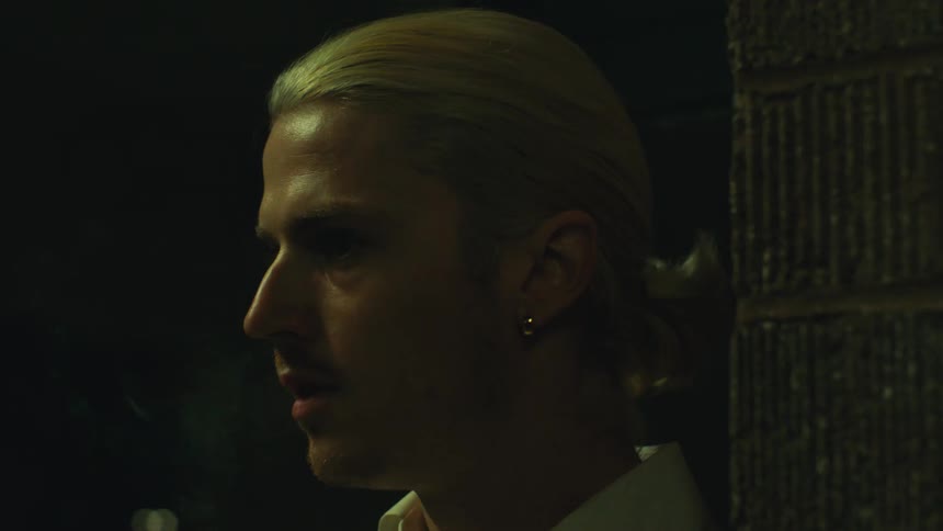 Portrait of a blond man in an outdoor setting next to a column at night smoking