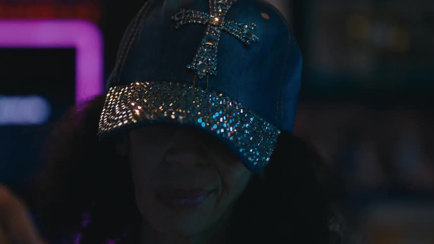 A woman with curly hair wearing a hiphop cap with a cross on it drinks a beverage in a bar