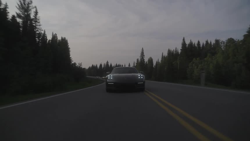 Line of vehicles drives along highway passing through a conifer forest under a cloudy sky
