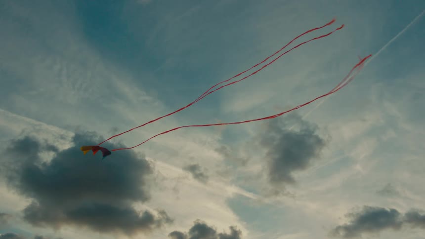 A kite with long red tails flies in the sky amidst scattered clouds during late afternoon