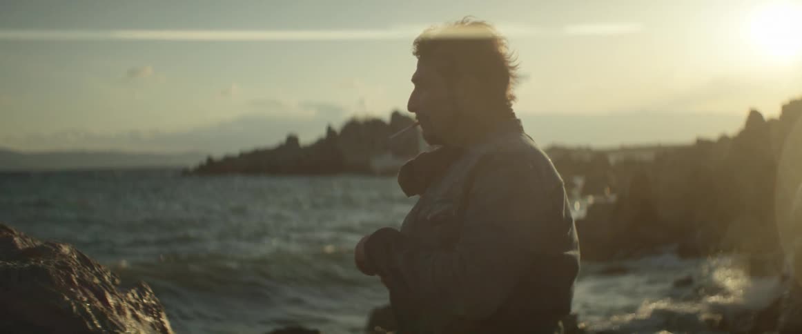 A man with a cigarette in his mouth bending over to light his cigarette as he stand next to the ocean on a windy day