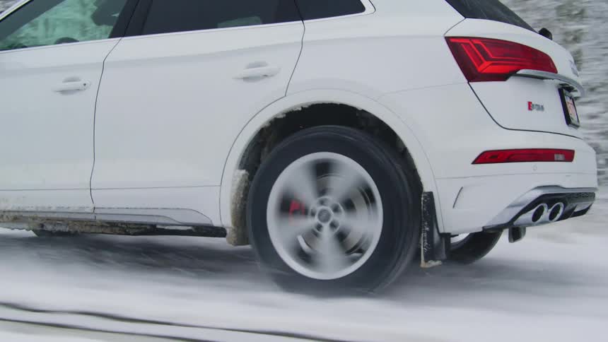 A closeup of a white cars driving through snowcovered ground near a forest
