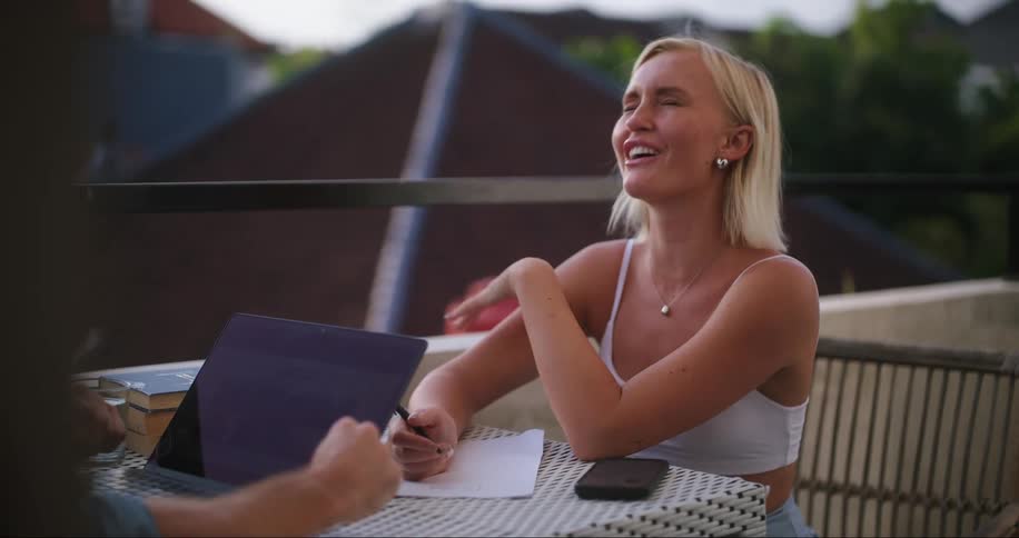 Woman laughs while working on balcony across a table from a man with a laptop