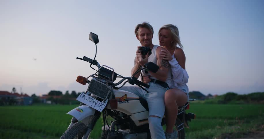 A young couple sitting on a motorcycle parked in the countryside taking pictures at dusk