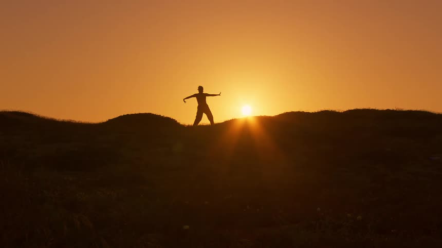 A silhouette of a person gesturing in contrast to the sun shining in the orange sky
