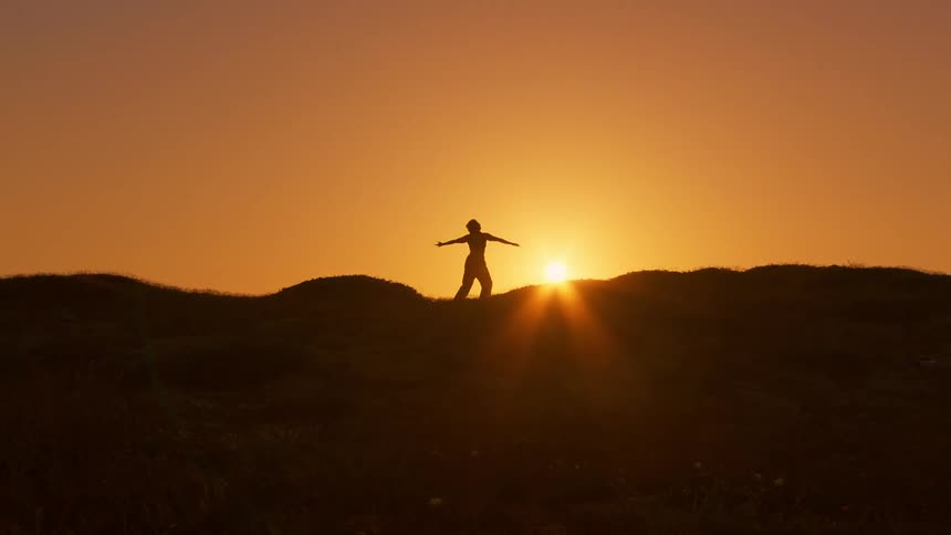 Person on top of a hill making body movements and in the background a sun on the golden horizon