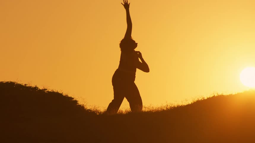 A silhouette of a woman dancing over a hill against the backdrop of a setting sun