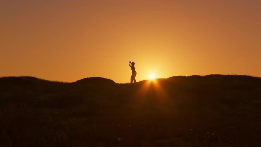 Silhouette of a mountain with a person on the peak dancing against a golden sun