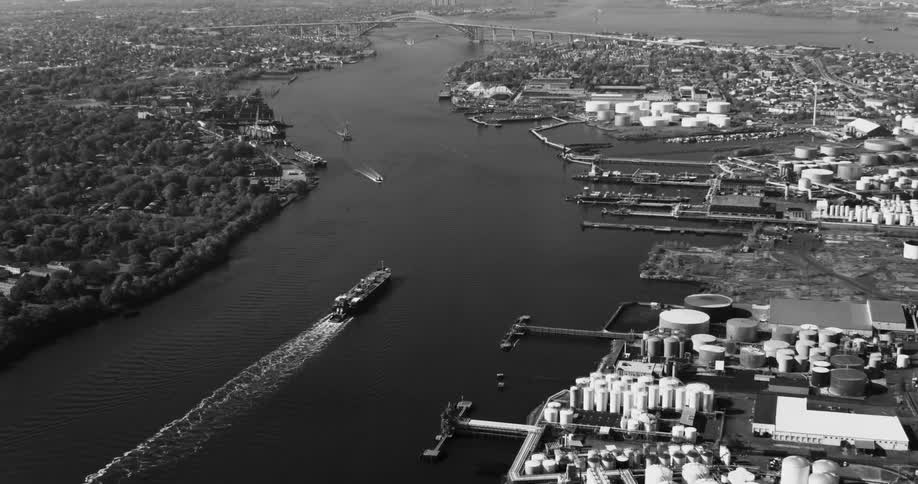 An aerial view of a large industrial area by a river with a ship creating ripples on the water