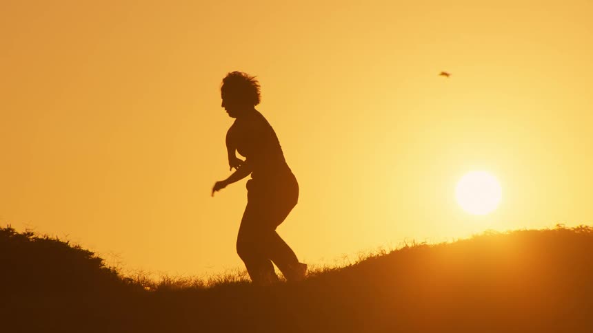 A silhouette of a woman dancing over a hill against the backdrop of a setting sun