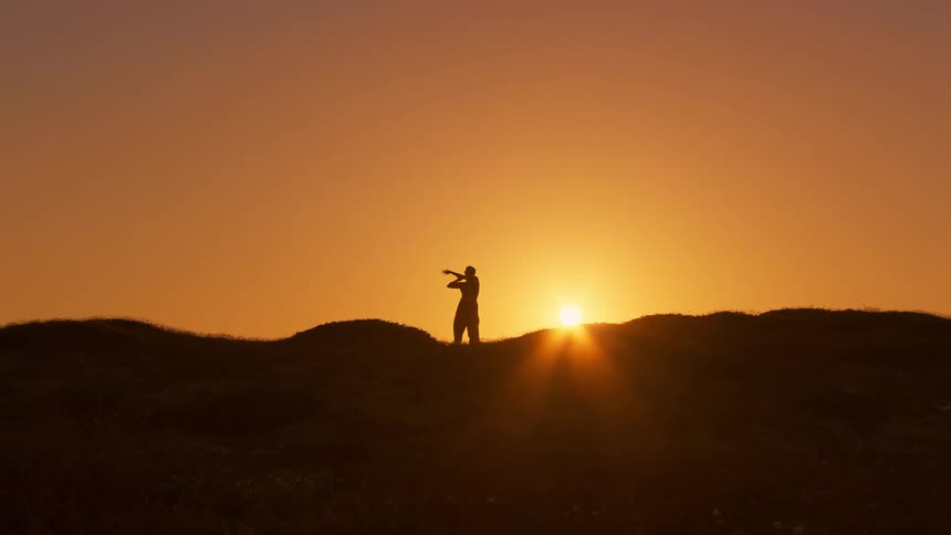 A silhouette of a woman dancing over a hill against the backdrop of a setting sun