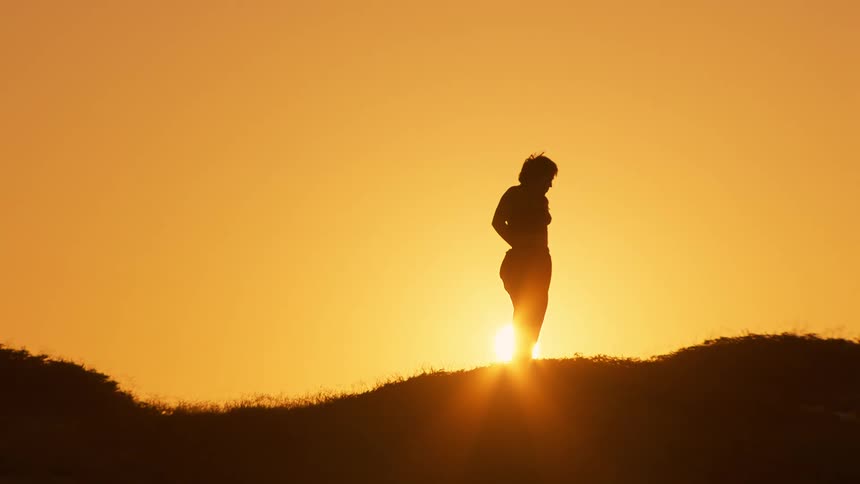 Silhouette of a woman stretching her body on a mountain peak against an orange sun