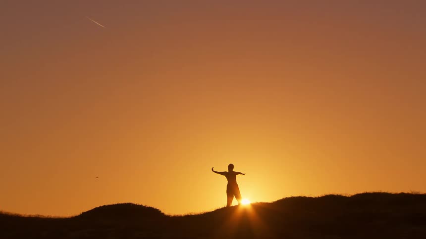 Silhouette of a person on top of a hill doing tai chi movements against the orange setting sun