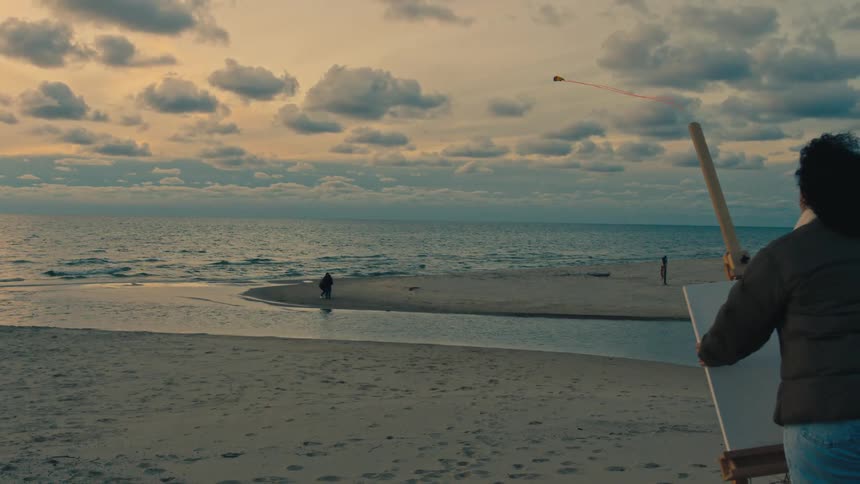 Female artist places canvas on easel and looks at people playing on the beach on a cloudy day