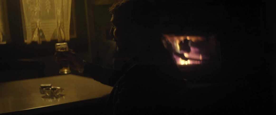 A man in black drinks a beer in front of a fireplace