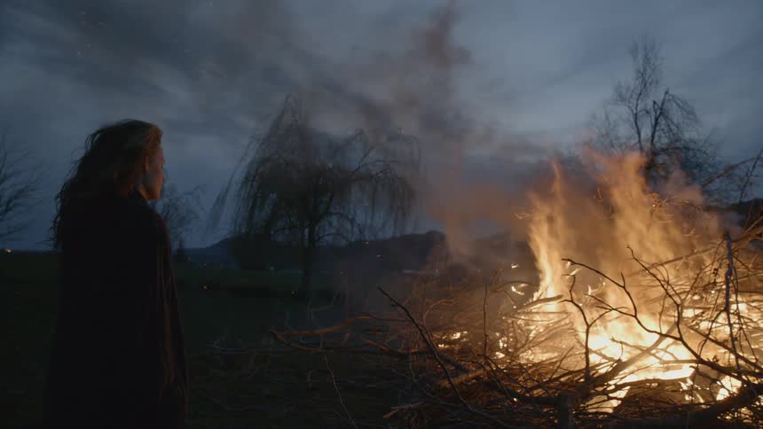 A woman stares into a fire burning a large pile of brush and branches