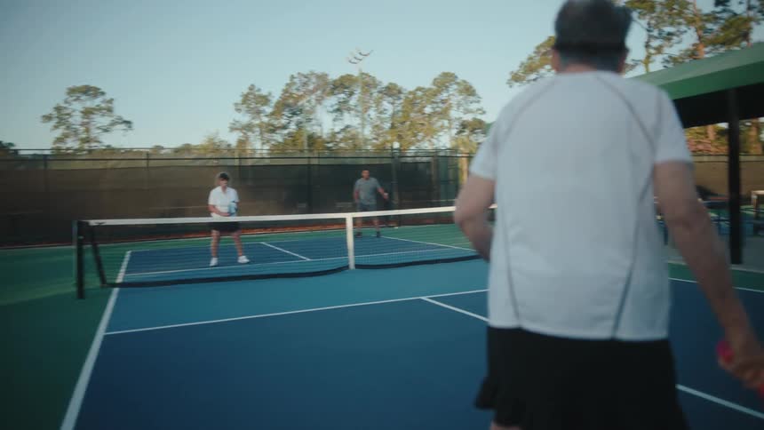 An older woman serves and charges the net in a game of pickleball