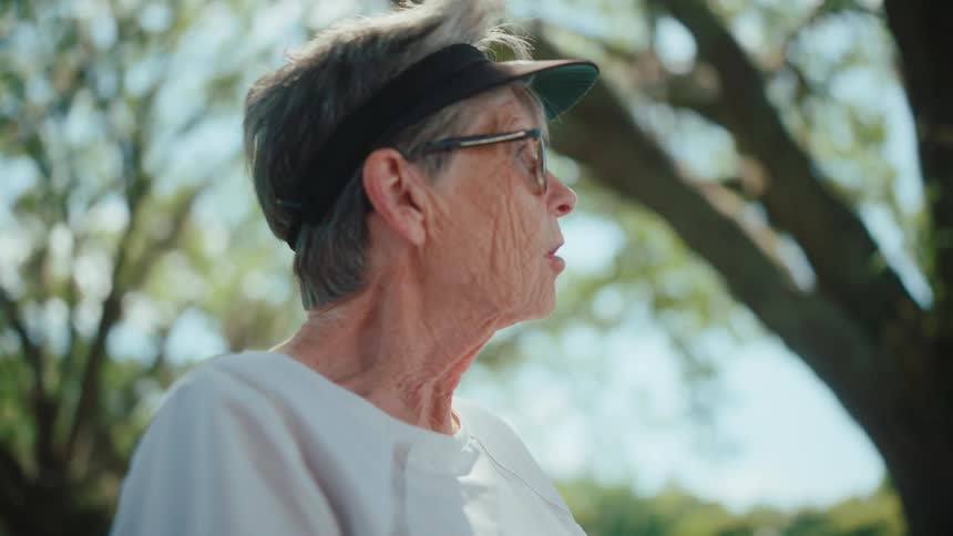 An elderly woman with glasses and a visor on her head is talking to someone next to her