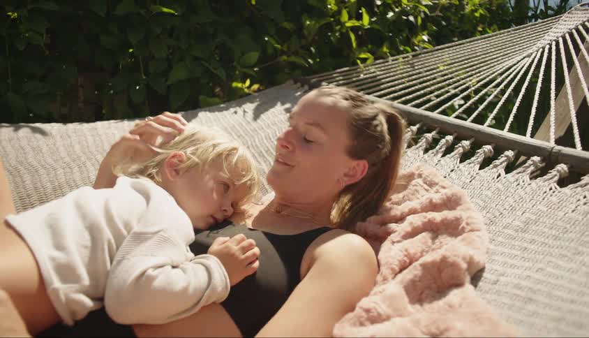A woman and her blond son are lying in a swing hammock and she gives her son a kiss and she is smiling