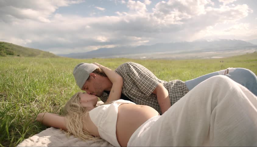 A man kisses a pregnant woman while they lie on a blanket in a field