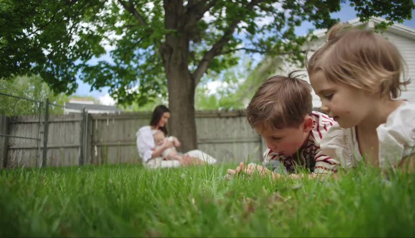Mother sitting with baby on her lap in a garden on the grass and two other children playing