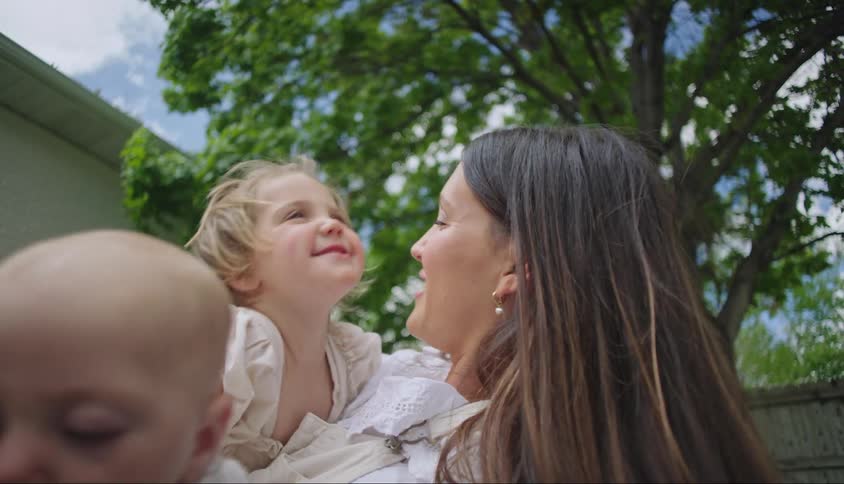 A mother lying in a yard with a baby laughs and hugs her toddler daughter