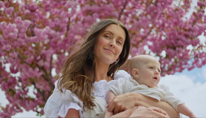 A mother holding her baby on her lap and there is a tree with pink leaves behind