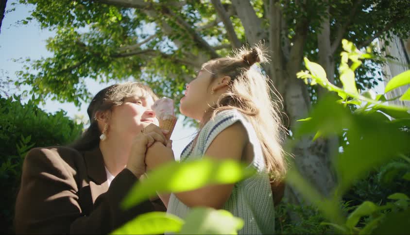 A mother licks her daughters ice cream cone while theyre in a park