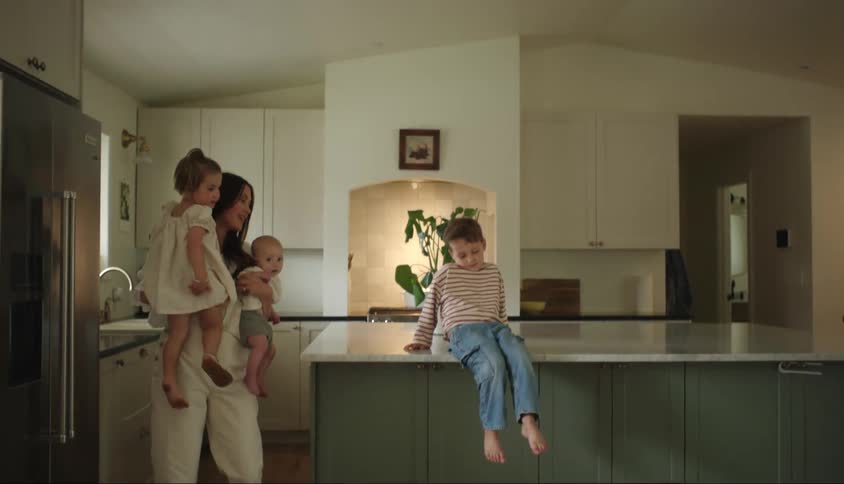 A mother carries two of her children while her son kicks his legs furiously in a kitchen