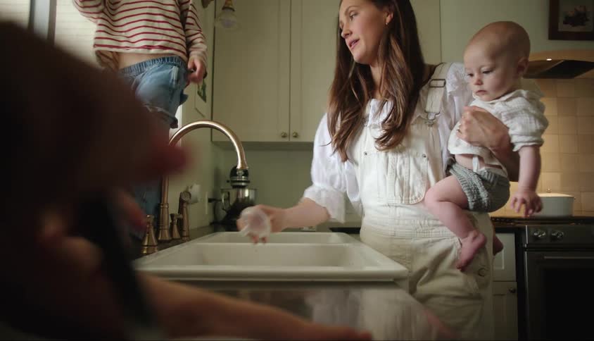 Mother holding a baby on her lap while standing in the kitchen at the sink and two children around