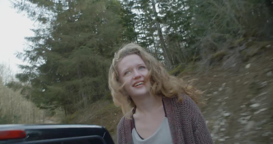 A woman rides in the back of an open bed truck going through a forest