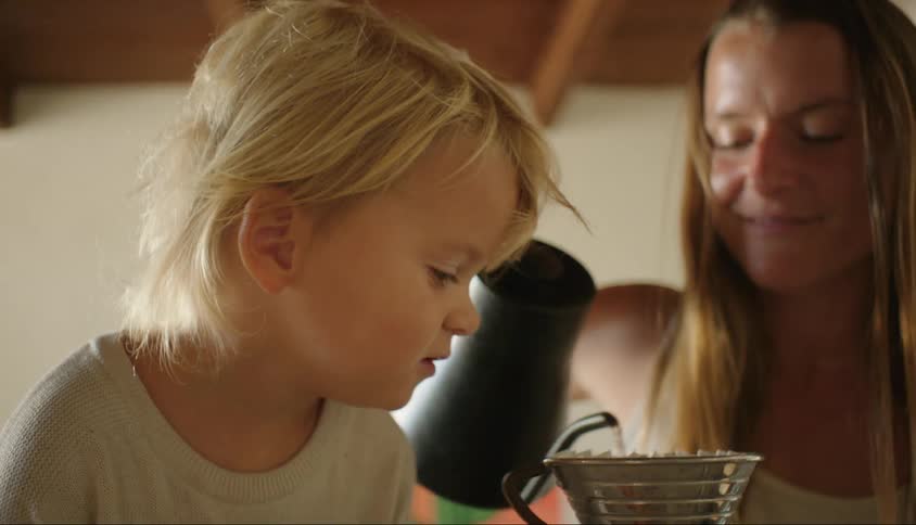 A toddler watches as his mother pours hot water into a coffee brewing device