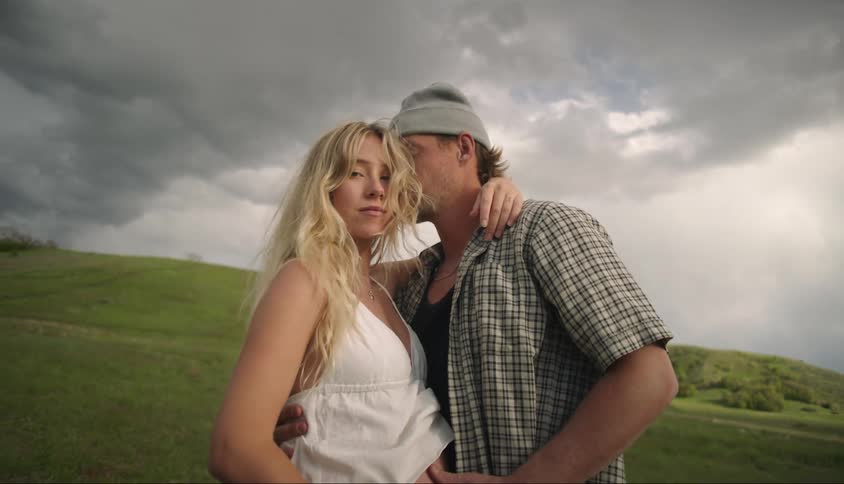 A man and a woman are hugging together and the woman is looking forward and behind is a green hill