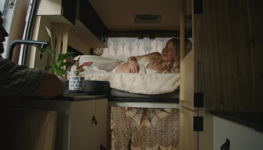 Woman lying on bed in moving camper rv rubs her pregnant belly while man sits near sink in cramped kitchen area