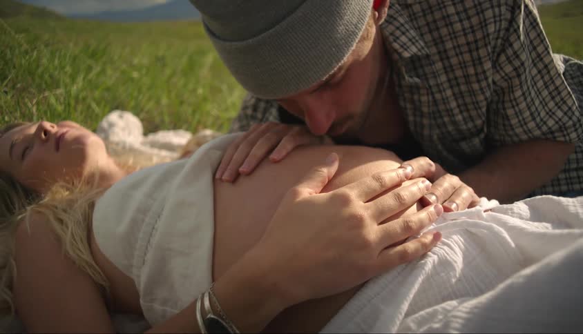 A man is kissing the belly of a pregnant woman who is lying on the grass
