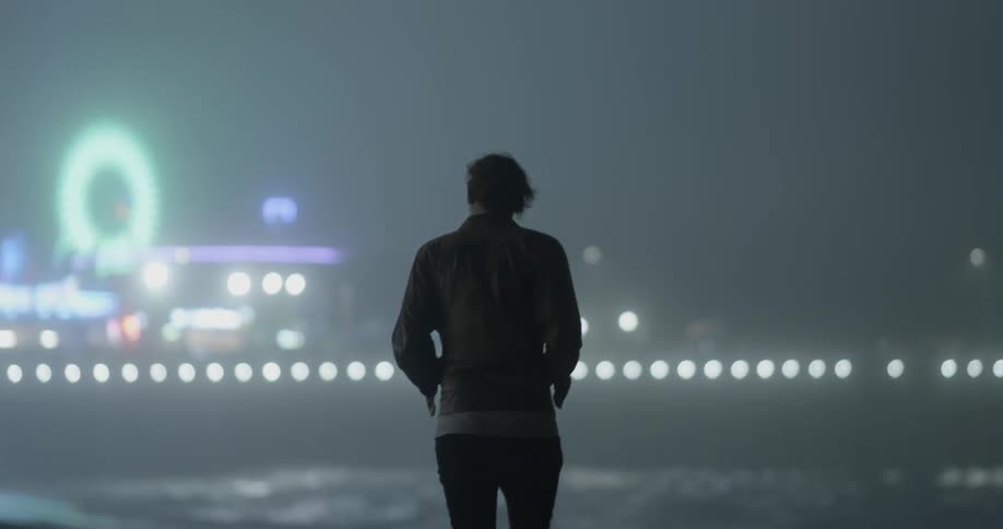 A man walks on the beach with shining lights in the distance