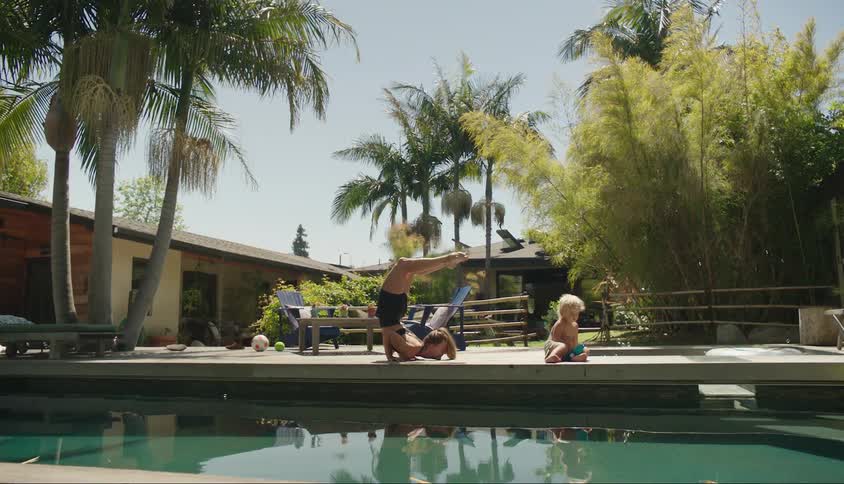 Woman stretching her body in an acrobatic way and with her little son playing by her side while they are by a swimming pool