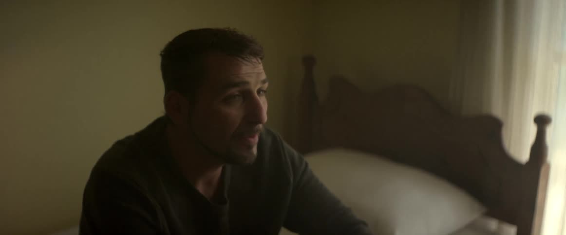 A man sits on a bed and smokes a cigarette as he talks