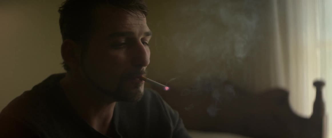 A man sits on a bed and lights a cigarette and shakes the ashes into a well used ashtray