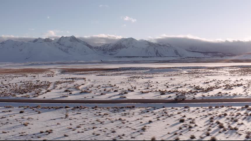 Aerial panorama of a car traveling along an extensive road with snowy mountains around