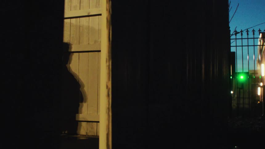 A silhouette of a person at a wooden door at night and on the right side there is a metal gate