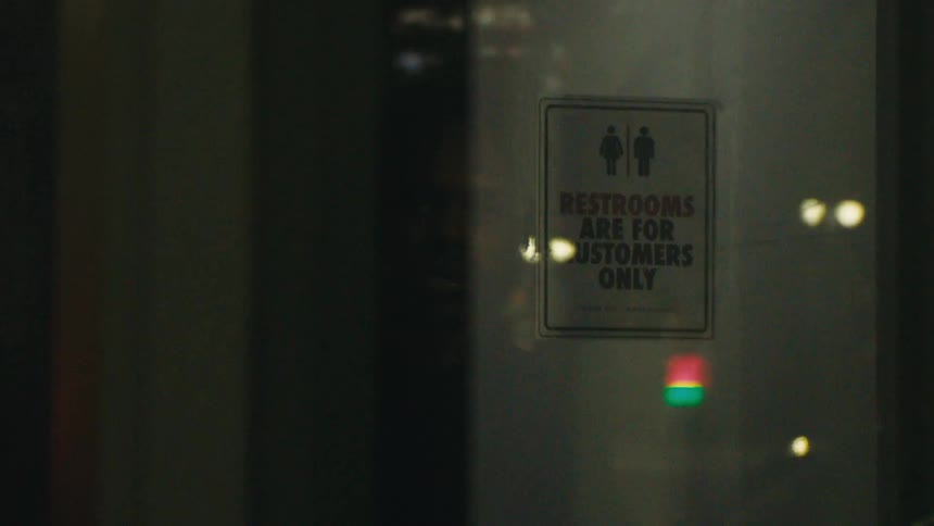 A restroom sign can be seen within a window that is reflecting automobiles lights