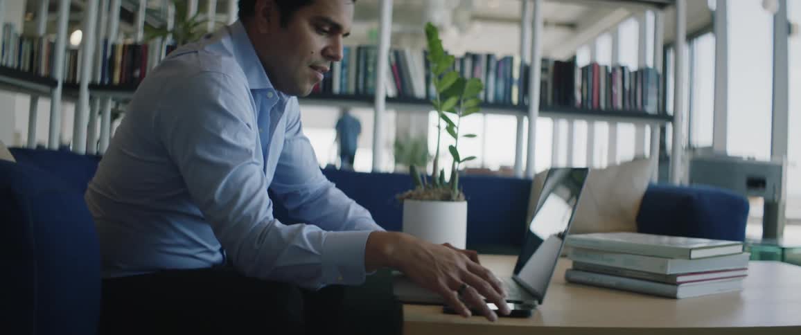 A man is sitting and using a cell phone on a table and next to it there is a laptop
