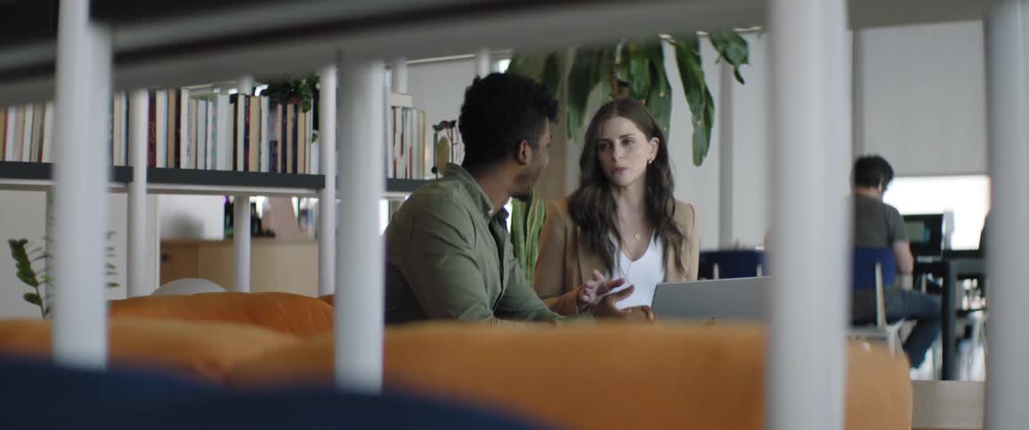 A woman and a man are sitting on a sofa while talking and there is a laptop in front of the woman